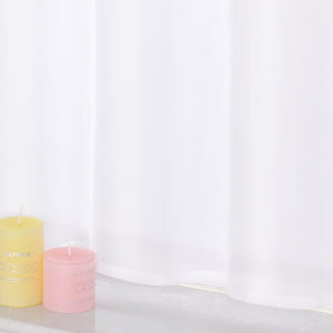 Der Vorhang ist aus leichtem, lichtdurchlässigem Material gefertigt. Auf der Fensterbank unter dem Vorhang stehen zwei Kerzen: eine gelbe und eine rosa, beide mit Text auf der Oberfläche.