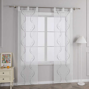 Joyswahl – Das Bild zeigt die Seitenansicht einer weissen transparenten Gardine mit Wellenperlenmuster und Stickerei, die an einer Metallstange haengt. Links vom Fenster steht ein heller Schubladenschrank mit einem farbigen Blumenbild und einem kleinen orangen Objekt darauf. Rechts vom Fenster steht eine Stehlampe mit einem weissen gestreiften Lampenschirm.