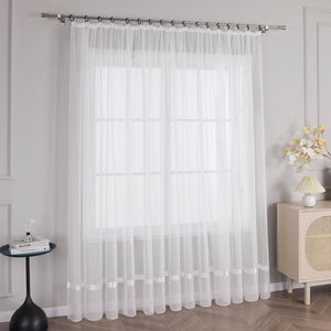 Vor der hellgrauen Wand befindet sich ein Fenster mit weißen Voile-Vorhängen mit Metallstangen mit schwarzen Ringen am oberen Ende. Links vom Fenster steht ein kleiner schwarzer runder Tisch mit ein paar Büchern und Vasen mit Blumen darauf. Die Vorhänge sind lang und hängen bis zum Boden, sie sind weiß und lichtdurchlässig.