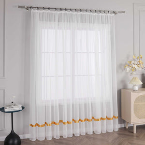 An einem Fenster haengt ein weisser Vorhang mit orangefarbenen Satinbaendern und Kraeuseln. Am oberen Ende befindet sich eine Metallstange mit schwarzen Ringen, die den Vorhang an seinem Platz halten. Auf der linken Seite des Fensters steht ein kleiner schwarzer runder Tisch mit einigen Gegenstaenden. Rechts vom Fenster steht ein heller Schrank mit einer Lampe mit hellem Schirm und einem Strauss dekorativer Blumen.
