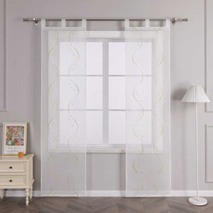 Vor einer hellgrauen Wand befindet sich ein Fenster, an dem eine weisse transparente Schiebegardine mit symmetrischem Wellenperlenmuster und grüner Stickerei haengt. Die Gardine ist an einer Metallstange mit Ringen befestigt. Links vom Fenster steht ein heller Schrank mit Schubladen, auf dem ein farbenfrohes Blumenbild und ein kleines oranges Objekt platziert sind. Rechts vom Fenster befindet sich eine Stehlampe mit weissem gestreiftem Lampenschirm.