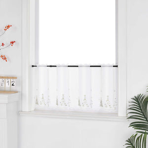 Eine weiße, transparente Scheibengardine mit goldenen Tannenbaum-Motiven hängt vor einem weißen Fenster. Die Wand ist ebenfalls weiß, was den hellen und festlichen Charakter unterstreicht. Seitlich sind dekorative Blumenarrangements zu sehen, die dem Bild eine natürliche und stimmungsvolle Note verleihen – ideal als stimmige Weihnachtsdekoration.