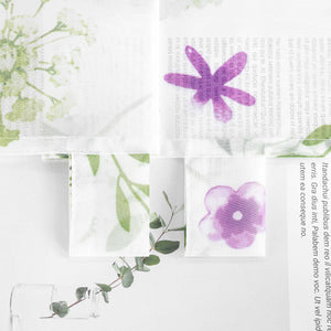 Ein Stoff mit Pflanzen- und Blumenmuster, darunter lila Blumen und grüne Zweige. Rechts steht ein transparenter Glasvase mit Eukalyptuszweigen.
