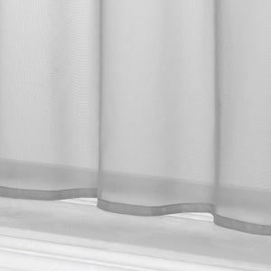 Dies ist eine Detailaufnahme des unteren Teils des grauen kurzen Vorhangs. Der Vorhang zeigt das leichte, halbtransparente Material sowie den wellenförmigen Saum am unteren Rand. Darunter ist die Fensterbank zu sehen.