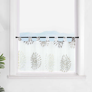 Die transparente Scheibengardine mit braunen Feuerwerksmotiven hängt elegant vor einem Fenster. Die weiße Wand im Hintergrund lässt die braunen Muster des Feuerwerks besonders hervorstechen und sorgt für eine ruhige, festliche Atmosphäre im Raum.