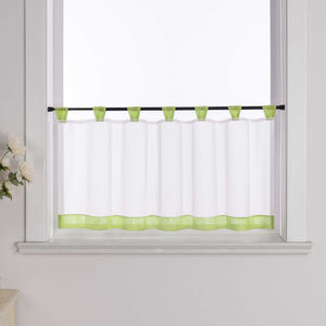 Die gruene Scheibengardine hängt vor einem Fenster, wobei die Hintergrundwand weiß ist. Der Vorhang ist transparent und wird von blumigen Dekorationen an der Seite begleitet.