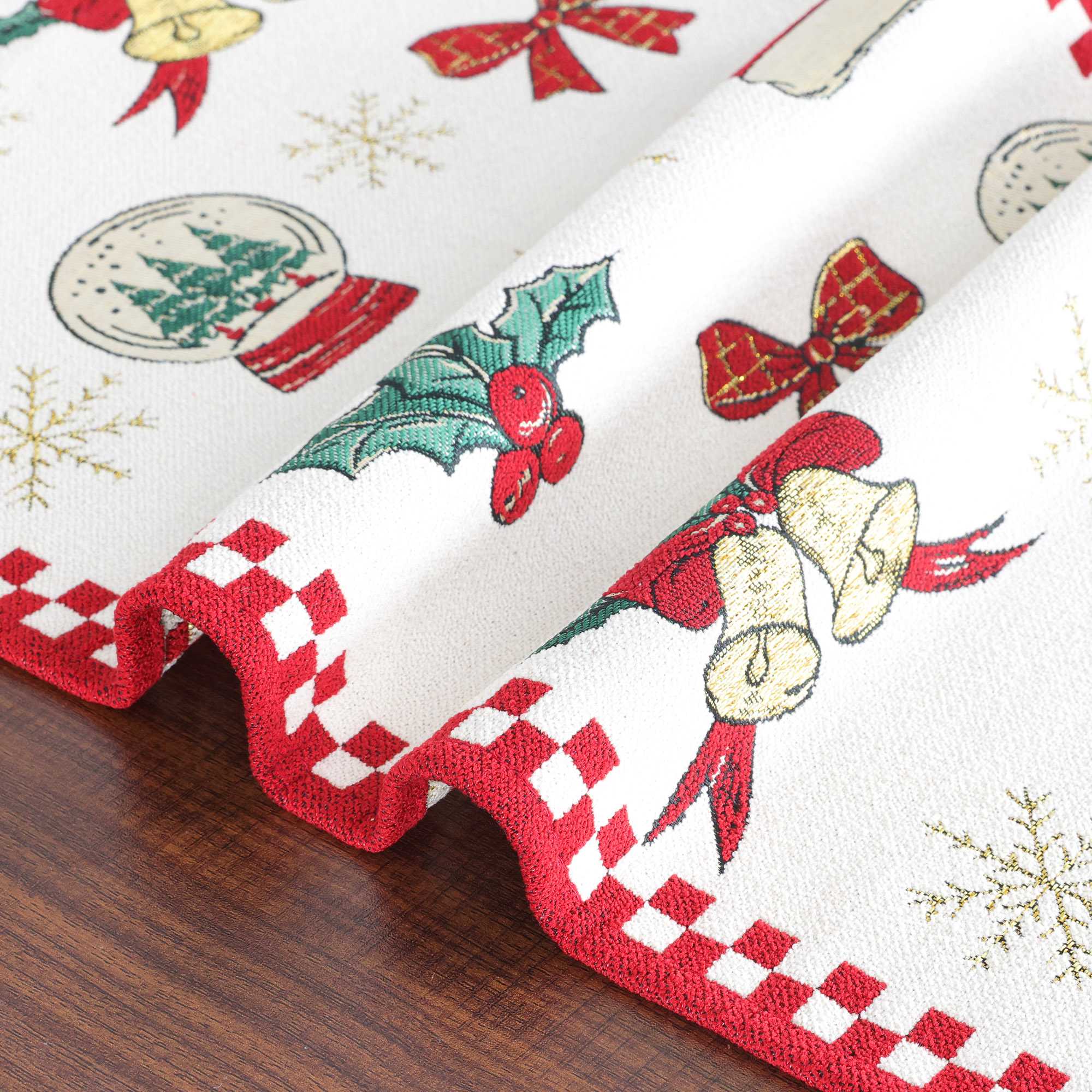 Dieses Bild zeigt die feinen Details eines Weihnachts-Tischläufers mit klassischem, festlichem Design. Die Ecken des Tischläufers sind sanft gefaltet, sodass der rote, karierte Rand und die weihnachtlichen Motive wie Glocken, Weihnachtskugeln und Schleifen gut zur Geltung kommen. Der Tischläufer kombiniert hochwertige Webtechnik und festliche Symbole, was ihn zu einem idealen Dekorationsstück für den Esstisch in der Weihnachtszeit macht.