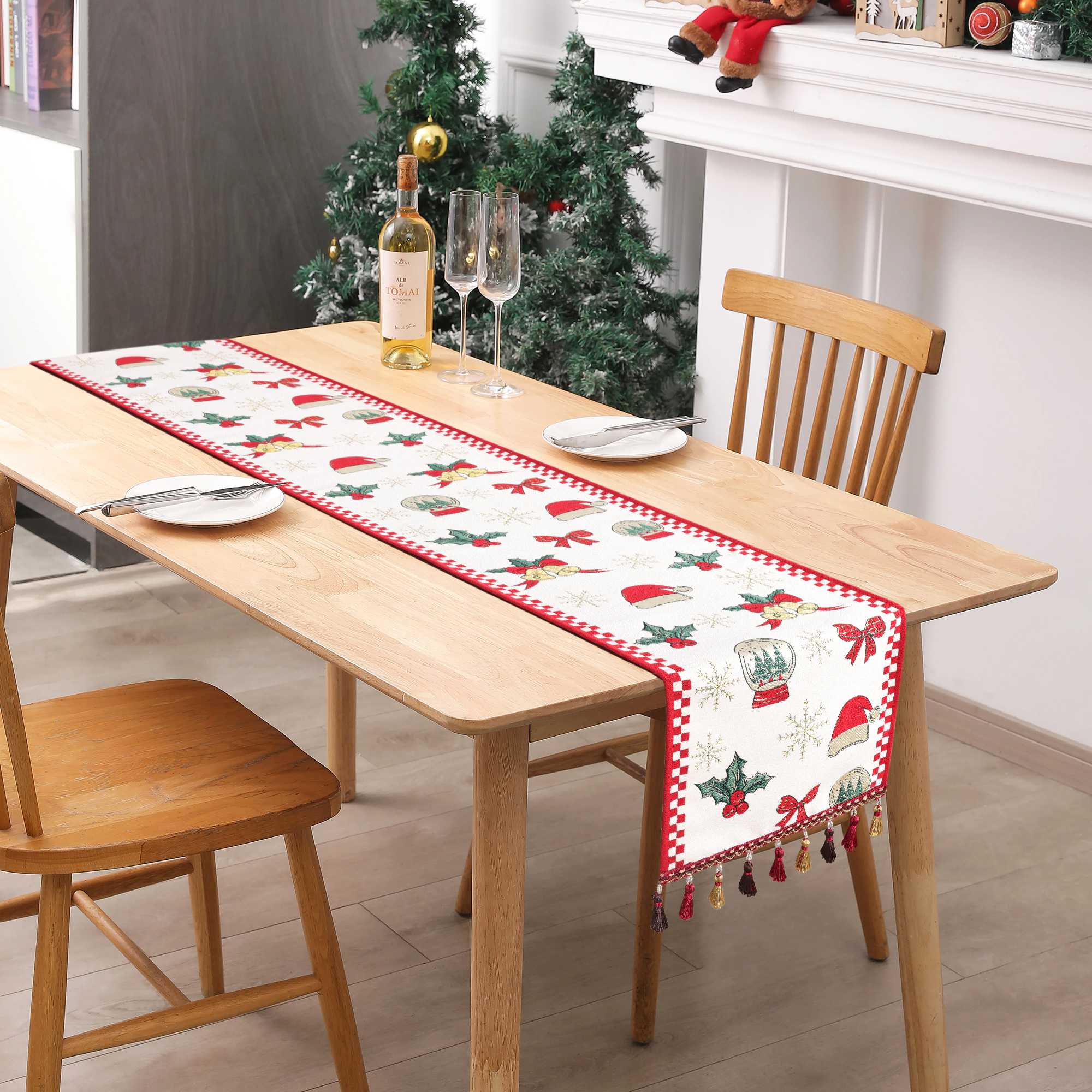 Dieses Bild zeigt einen Weihnachts-Tischläufer, der auf einem Esstisch platziert ist, umgeben von einer festlich dekorierten Weihnachtsbaum und weiteren Dekorationen. Der Tischläufer ist mit klassischen Weihnachtsmotiven wie Weihnachtsmützen, Glocken, Tannenbäumen und roten Schleifen verziert, die eine warme und festliche Atmosphäre schaffen. Die edlen Fransen am Rand des Tischläufers verleihen ihm zusätzlichen Charme und Eleganz, wodurch er sich perfekt für Weihnachtsfeiern und als Dekoration für den Essti