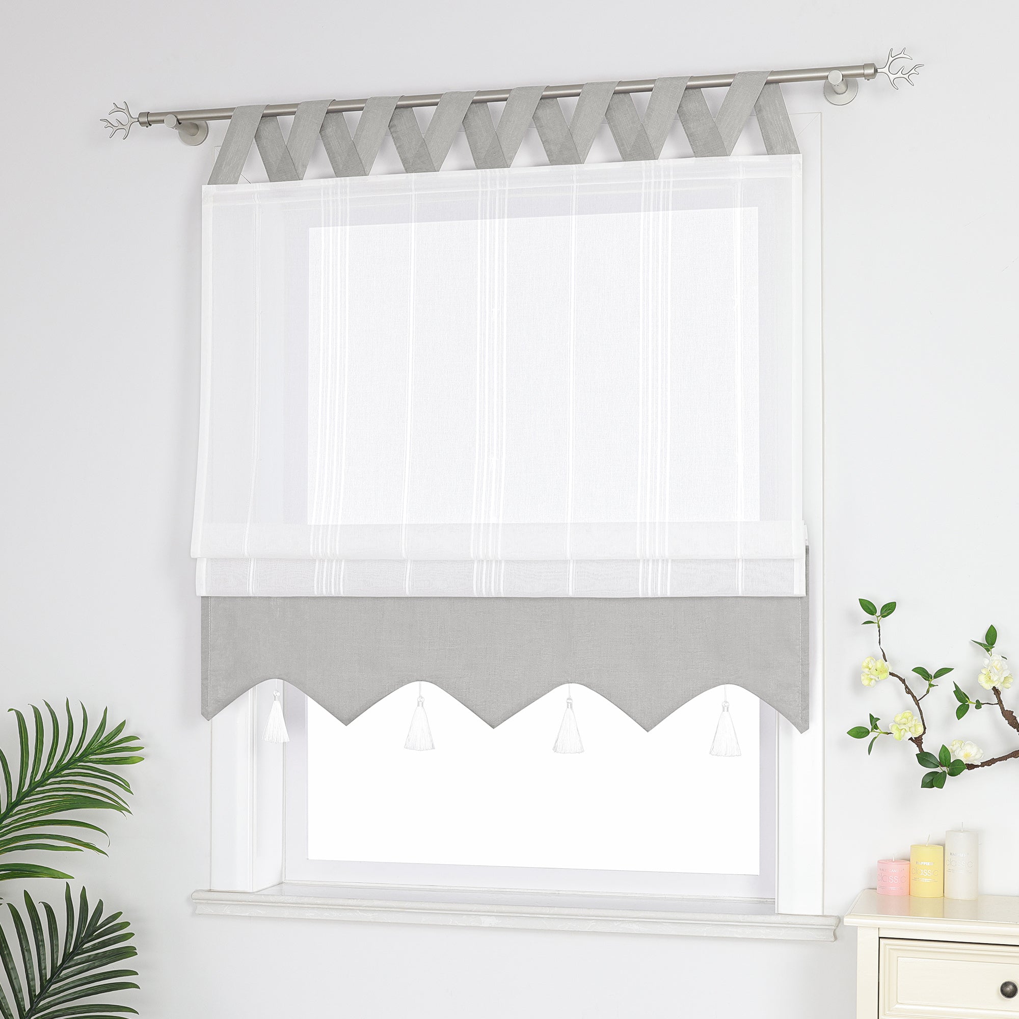 Graue und weisse Raffrollo Gardine mit Schlaufen dekorativen Quasten und geschwungenem Saum halb hochgezogen, Seitenansicht eines weißen Raffrollos mit Quasten und Schlaufen – halb geschlossen, sodass die Fenster nicht vollständig bedeckt sind, neben Grünpflanzen und duftenden Ornamenten.