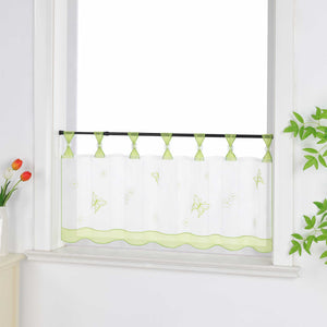 Seitliche Ansicht eines gruenen transparenten Kurzgardine mit dekorativen Perlen an den Aufhängeschlaufen und Schmetterlingsmuster auf der Oberfläche. Der Vorhang hat ein gruenes Dekoband am unteren Rand und ist vor einem Fenster mit einem Schrank und Blumen als Dekoration aufgehängt.