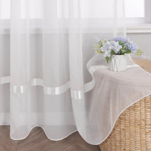 Am Fenster haengt ein weisser Vorhang aus Voile, auf dem sich mehrere weisse Dekobaender befinden. Rechts neben dem Fenster steht ein Objekt aus Flechtmaterial, das mit einem hellbeigen Stoff bedeckt ist. Darauf steht eine weisse Vase mit einem Strauss aus blau-weissen Blumen.