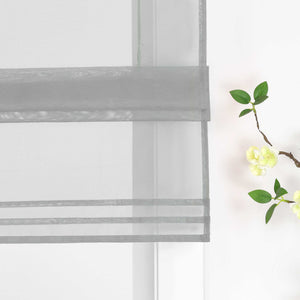 Detailansicht des unteren Bereichs eines halbtransparenten grauen Raffrollos. Der Stoff ist sanft hochgezogen und bildet dekorative Falten. Eine stilvolle Blumen-Deko neben dem Fenster rundet das Gesamtbild harmonisch ab.