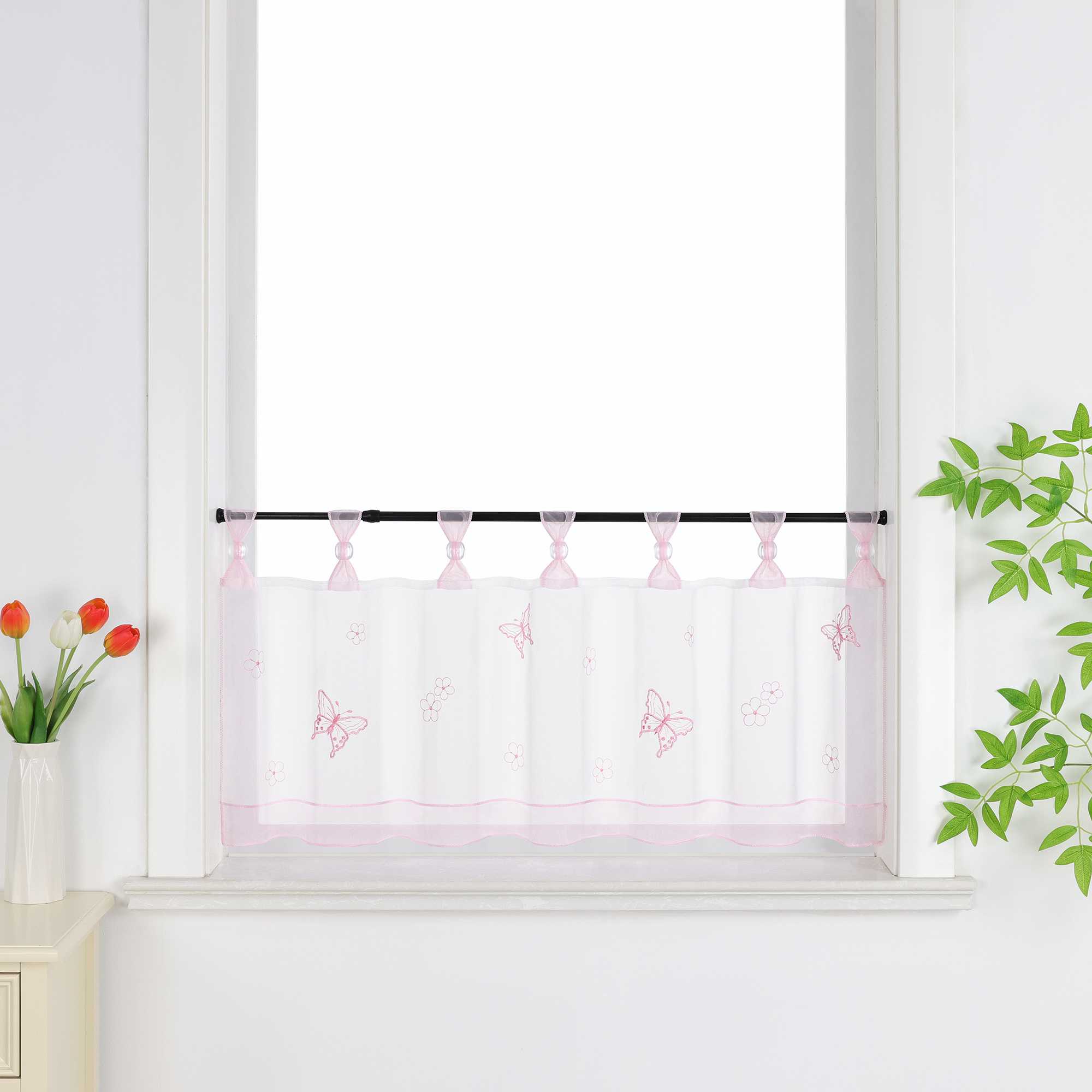 Rosa transparenter Kurzvorhang mit dekorativen Perlen und Schmetterlingsmuster, verziert mit einem rosa Dekoband am unteren Rand, aufgehängt neben einem Fenster mit Blumen und einem Schrank.