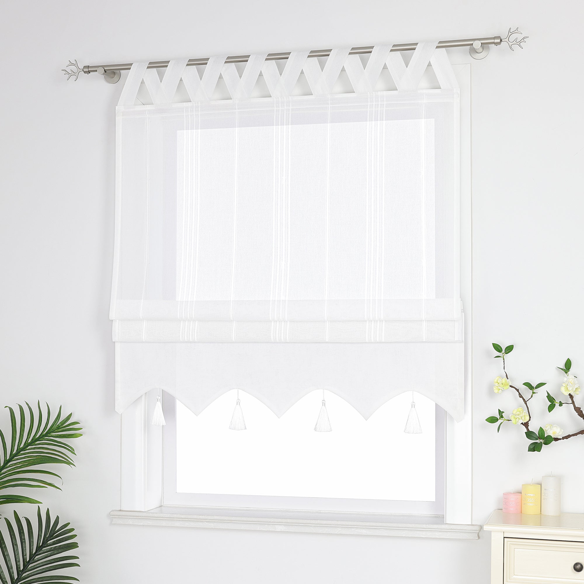 Seitenansicht eines weißen Raffrollos mit Quasten und Schlaufen – halb geschlossen, sodass die Fenster nicht vollständig bedeckt sind, neben Grünpflanzen und duftenden Ornamenten.