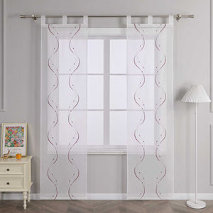 Vor einer hellgrauen Wand befindet sich ein Fenster, an dem eine weisse transparente Schiebegardine haengt. Die Gardine ist mit symmetrischen lila wellenfoermigen Perlenmustern und Stickereien versehen. Sie ist an einer Metallstange mit Schlaufen befestigt. Links vom Fenster steht ein heller Schrank mit Schubladen, auf dem ein farbenfrohes Blumenbild und ein orangefarbenes Objekt platziert sind. Rechts vom Fenster steht eine Stehlampe mit einem weiss gestreiften Lampenschirm.