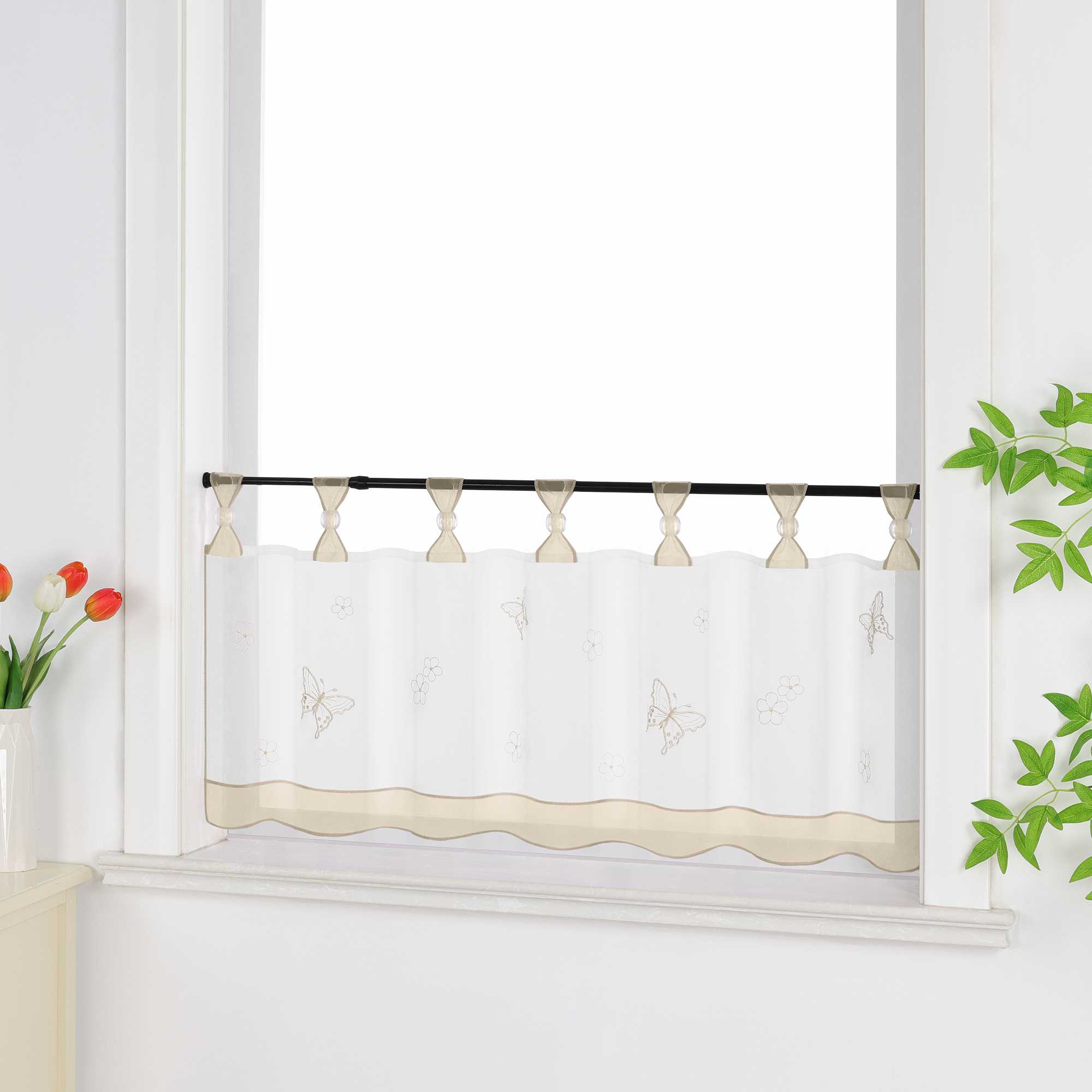 Seitliche Ansicht eines beige transparenten Kurzvorhangs mit dekorativen Perlen an den Aufhängeschlaufen und Schmetterlingsmuster auf der Oberfläche. Der Vorhang hat ein beiges Dekoband am unteren Rand und ist vor einem Fenster mit einem Schrank und Blumen als Dekoration aufgehängt.