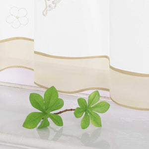 Nahaufnahme des unteren Teils eines beigen transparenten Vorhangs mit Schmetterlings- und kleinen Blumenmustern. Der Vorhang ist mit einem beigen Dekoband am unteren Rand verziert.