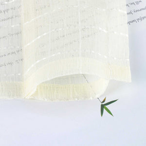 Der cremeweiße Vorhangstoff liegt locker gebogen auf einer hellen Fläche. Die feine Karostruktur und luftige Transparenz sind deutlich erkennbar. Ein grünes Blatt unter dem Stoff betont die weiche Textur und Lichtdurchlässigkeit.