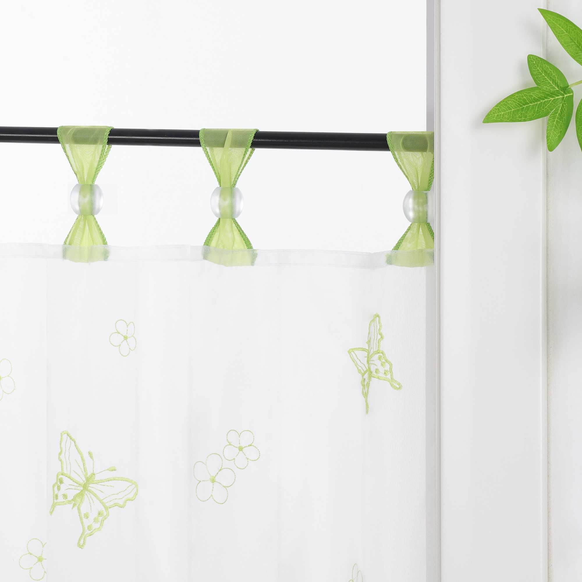 Nahaufnahme der gruenen Vorhangringe mit transparenten Perlen, der Vorhang hängt an einer Stange und zeigt Schmetterlingsmuster, daneben sind Blumen als Dekoration.
