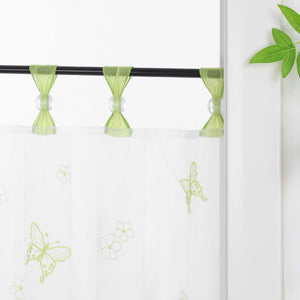 Nahaufnahme der gruenen Vorhangringe mit transparenten Perlen, der Vorhang hängt an einer Stange und zeigt Schmetterlingsmuster, daneben sind Blumen als Dekoration.