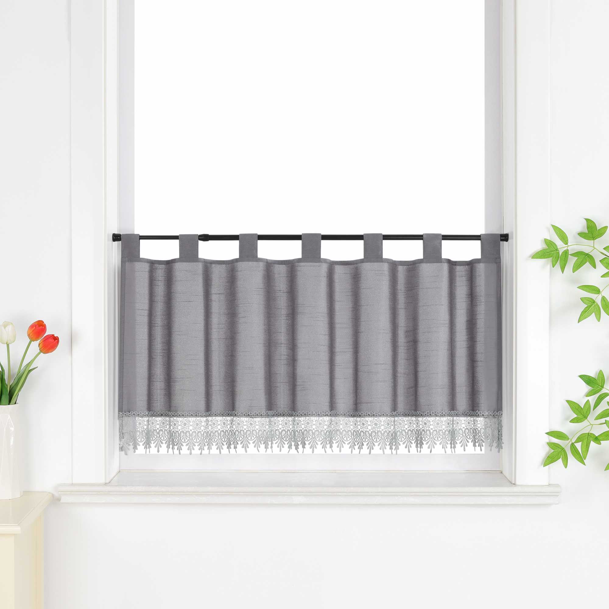 Graue Scheibengardine hängt vor einem Fenster mit weißer Wand. Daneben sorgen rote Blumen, grüne Blätter und eine Vase für eine frische und dekorative Note.