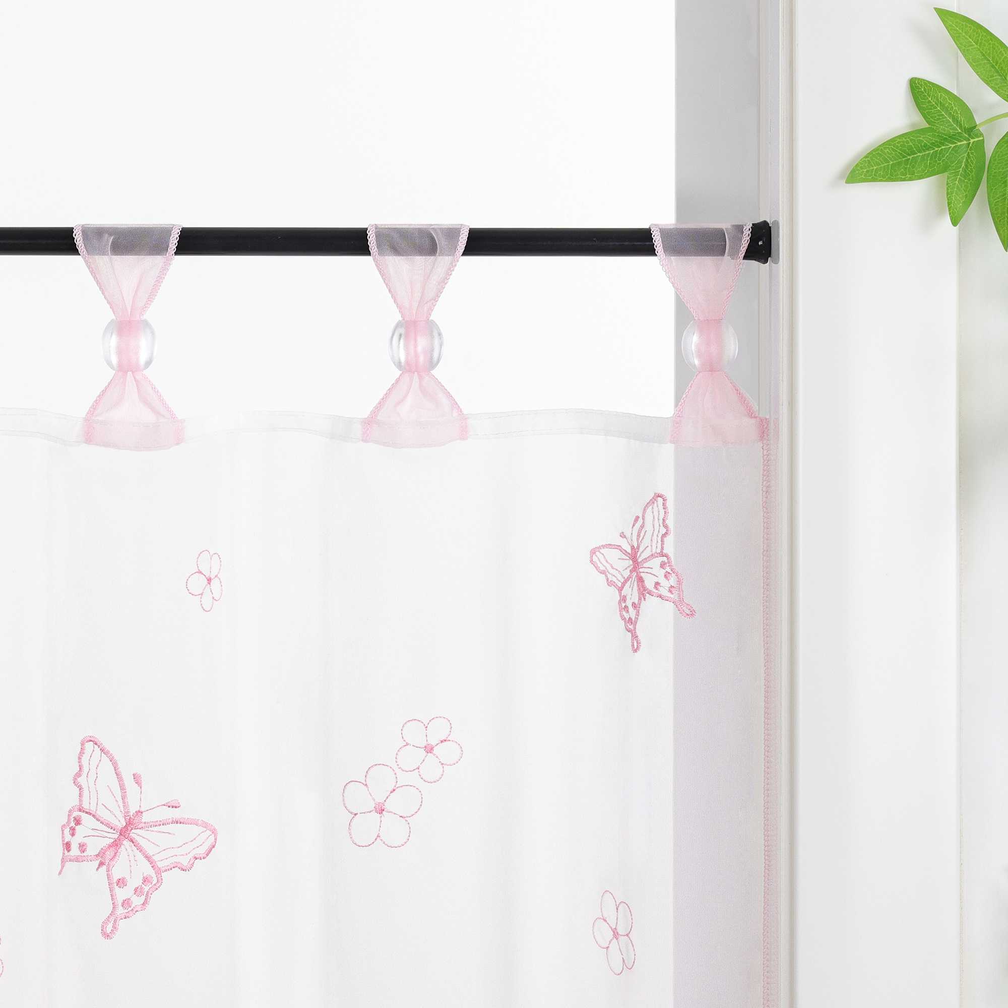 Nahaufnahme der rosa Vorhangringe mit transparenten Perlen, der Vorhang hängt an einer Stange und zeigt Schmetterlingsmuster, daneben sind Blumen als Dekoration.
